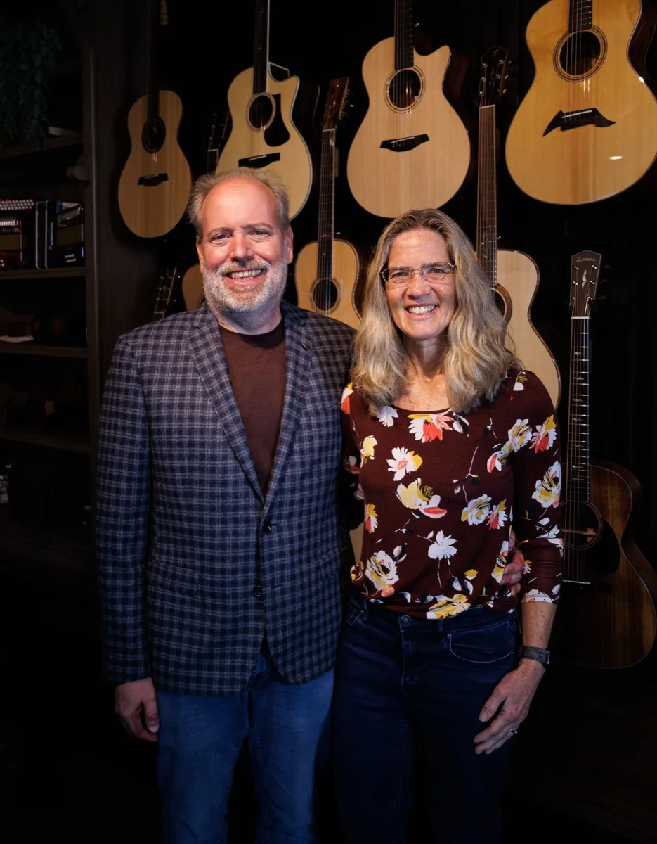 Ken and Laurie standing together in the Corzic Music showroom, in front of a wall of acoustic guitars